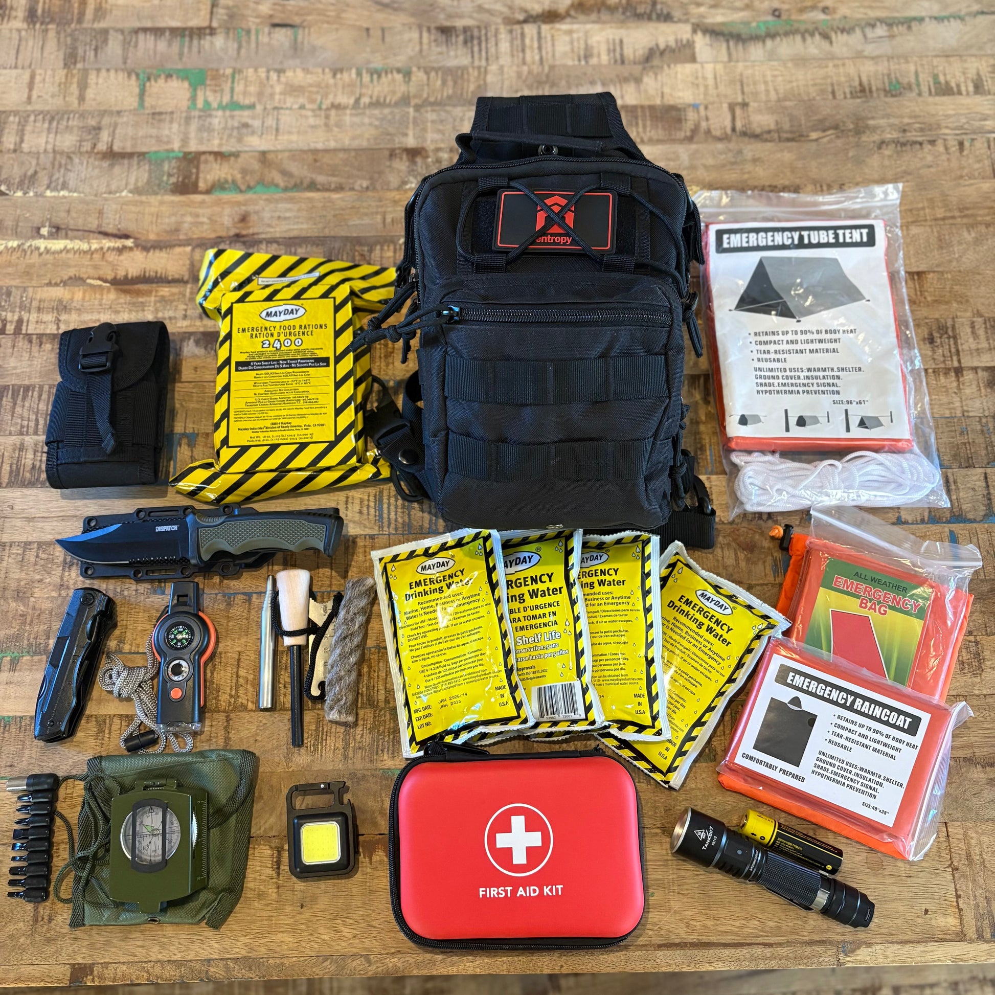 Collection of emergency supplies including a black backpack, first aid kit, and tools on a wooden surface.